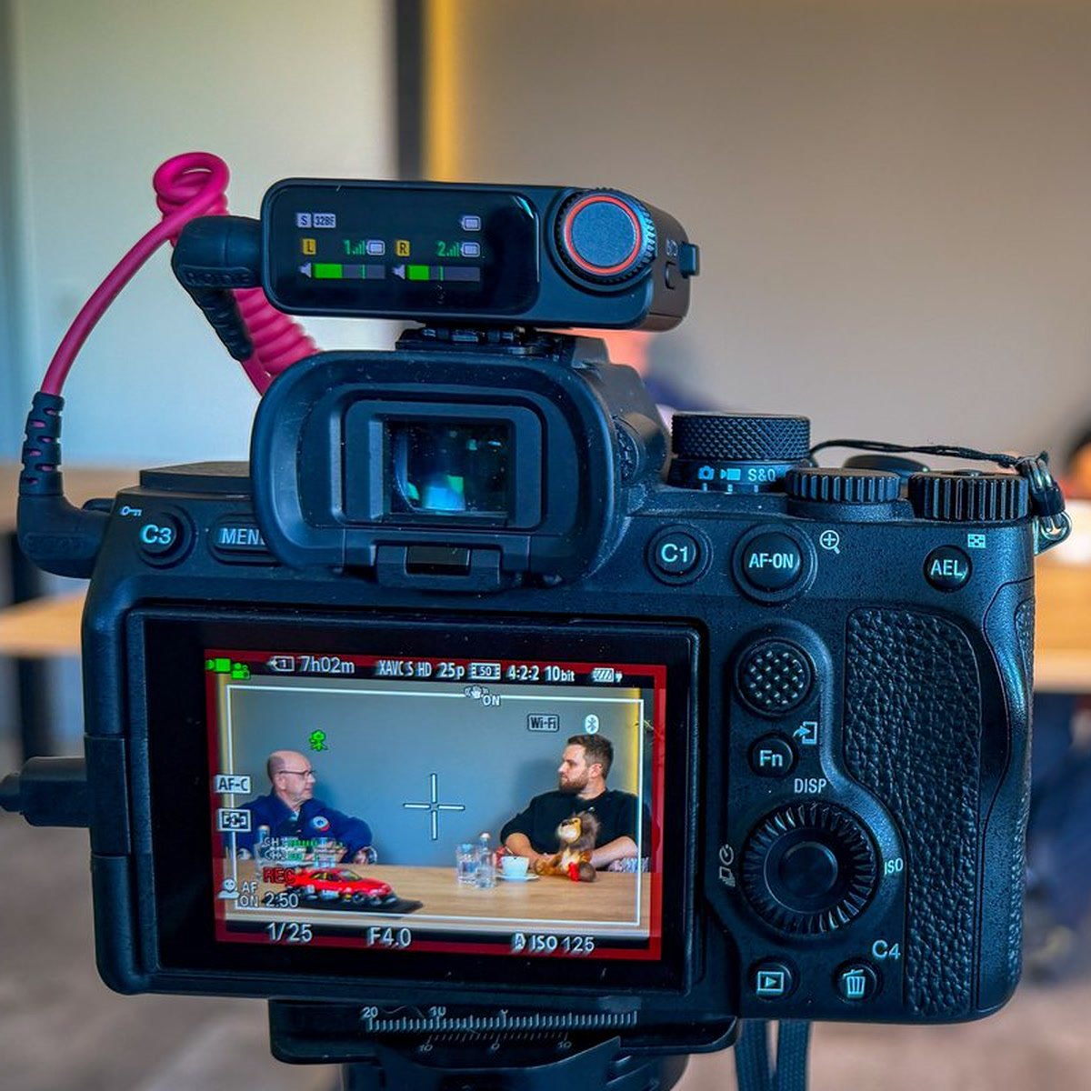 Kameradisplay bei einem Interviewdreh im Autohaus, auf dem Monitor zwei Gesprächspartner am Tisch mit einem roten Modellauto