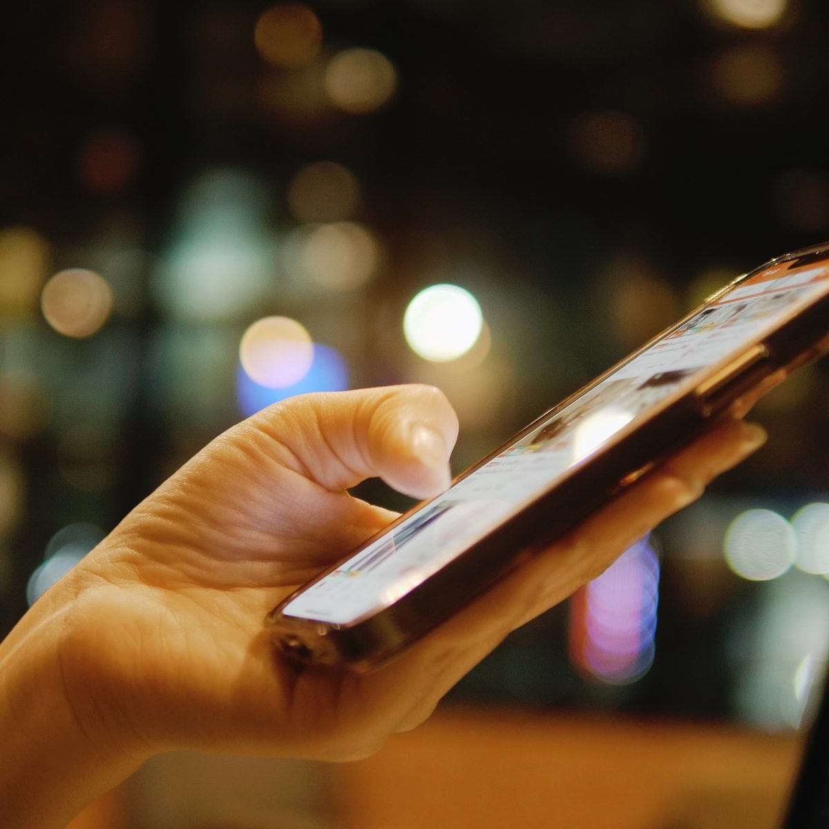 Hand hält abends ein Smartphone und scrollt durch einen Social-Media-Feed, im Hintergrund unscharfe Lichter